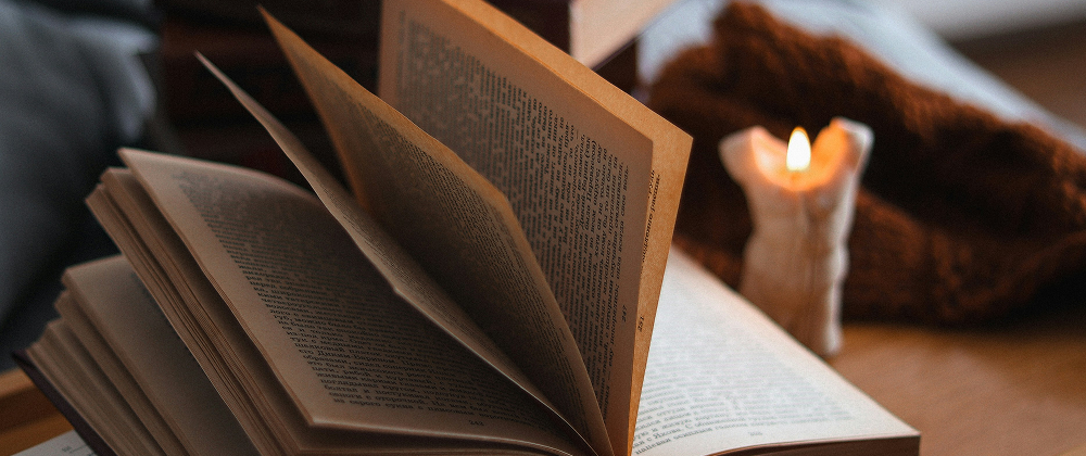 Open vintage book with turning pages beside a lit candle and stacked books on a wooden tray — cosy, warm reading atmosphere indoors.