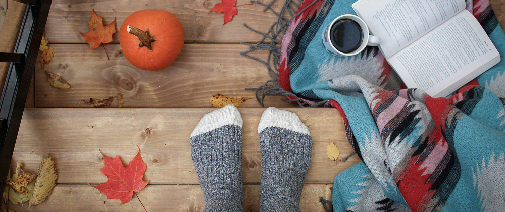 Cosy autumn scene with wool socks on a wooden deck, a pumpkin, colorful autumn leaves, open book, coffee mug, and patterned blanket.