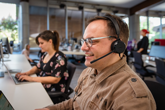 A man wearing glasses and a headset smiles while working on a computer in a modern open-plan office, with coworkers typing at nearby desks.