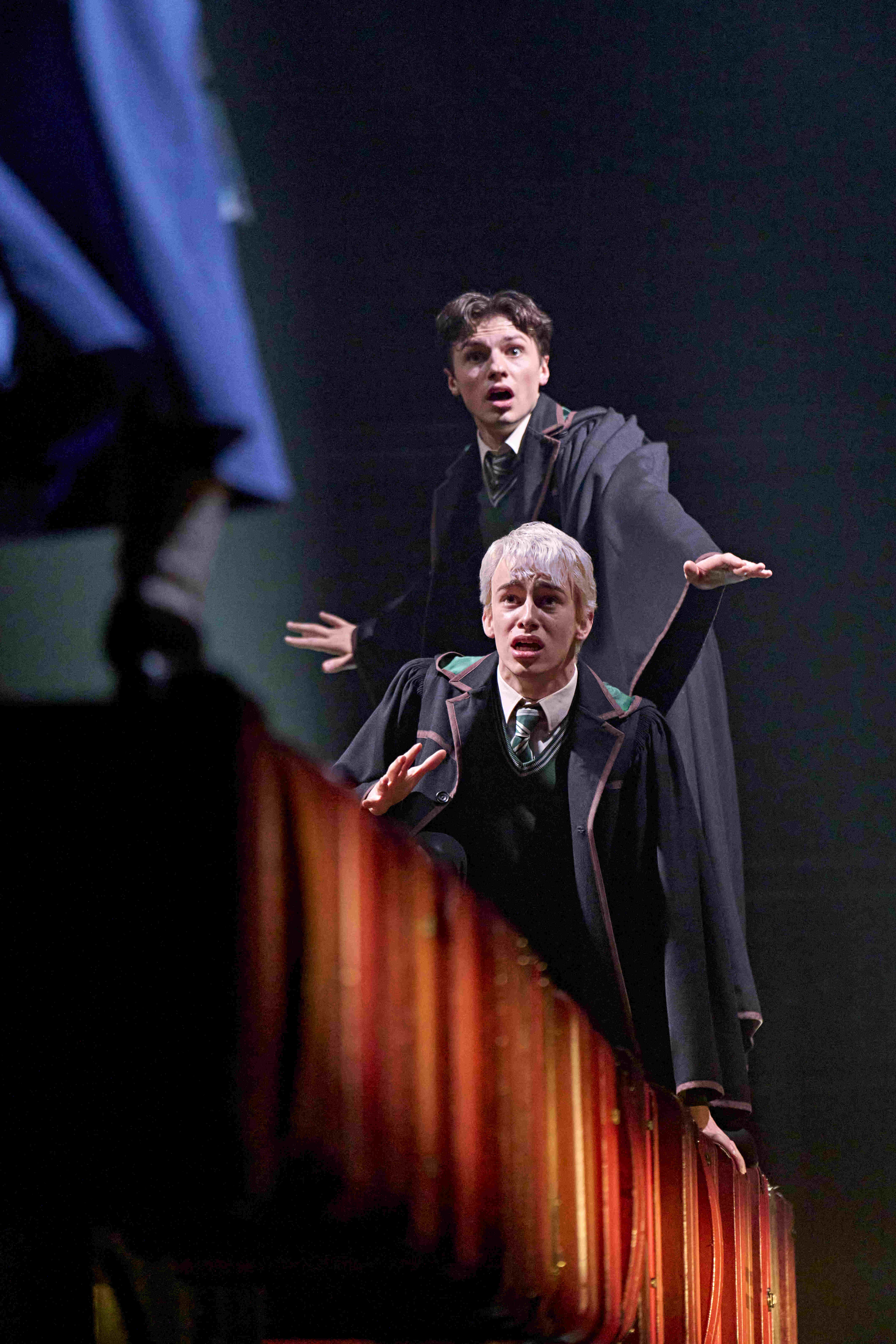 Characters examining a book in front of a large bookshelf, discussing a magical mystery, from Harry Potter and the Cursed Child.