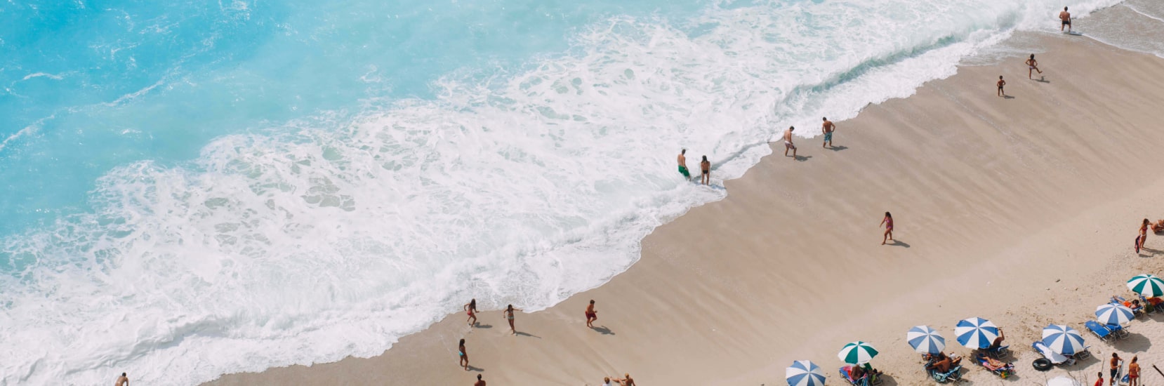Tourists relaxing under umbrellas and walking along a sandy beach with turquoise waves crashing on the shore.