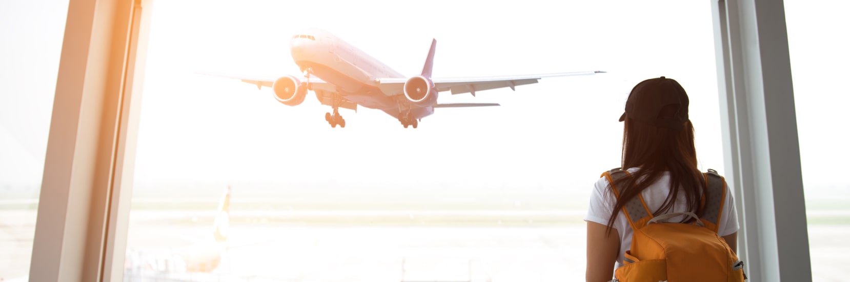 Female traveller with backpack watching airplane take off through airport window at sunrise.