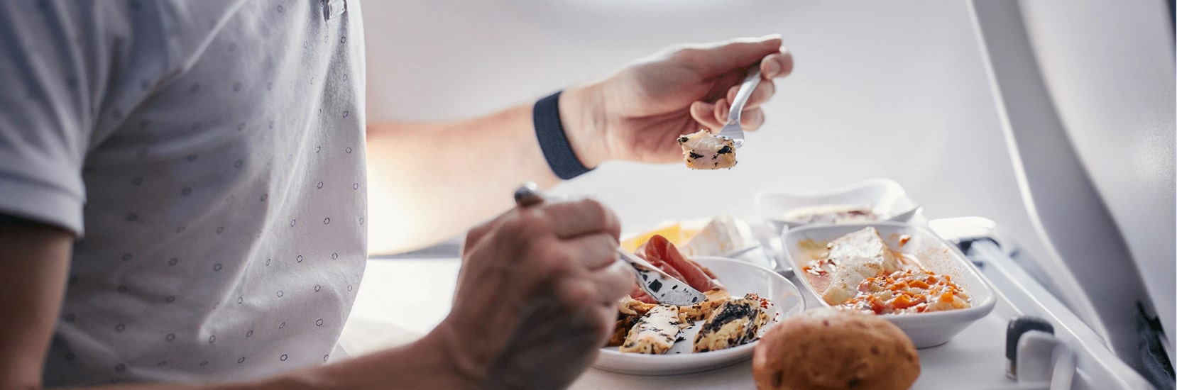 Passenger eating an in-flight meal served on a tray table, with dishes including chicken, salad, cheese, and bread