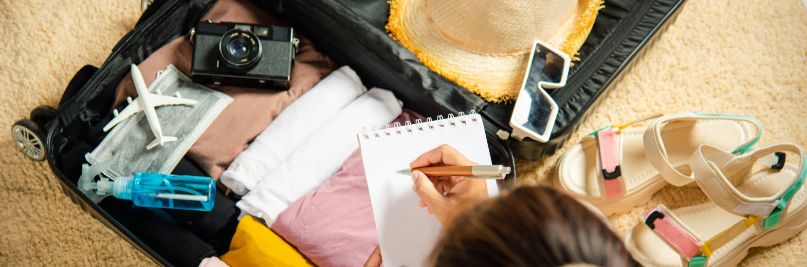 Under 18 Travel Insurance - An aerial view of a girl about to write in her notepad and packing her suitcase for a trip. Packed items include clothes, a camera, sunglasses and sandals to wear.