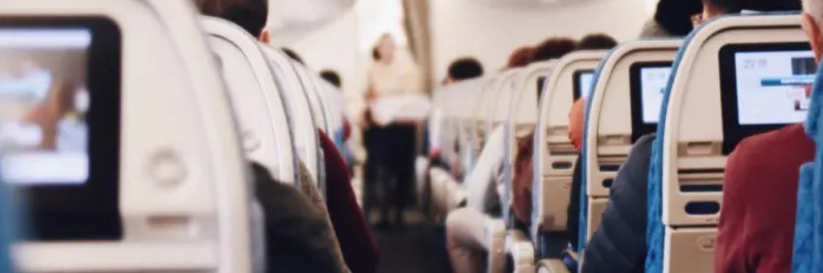 View down the aisle of an airplane cabin with rows of economy seats, seatback screens, and passengers seated.
