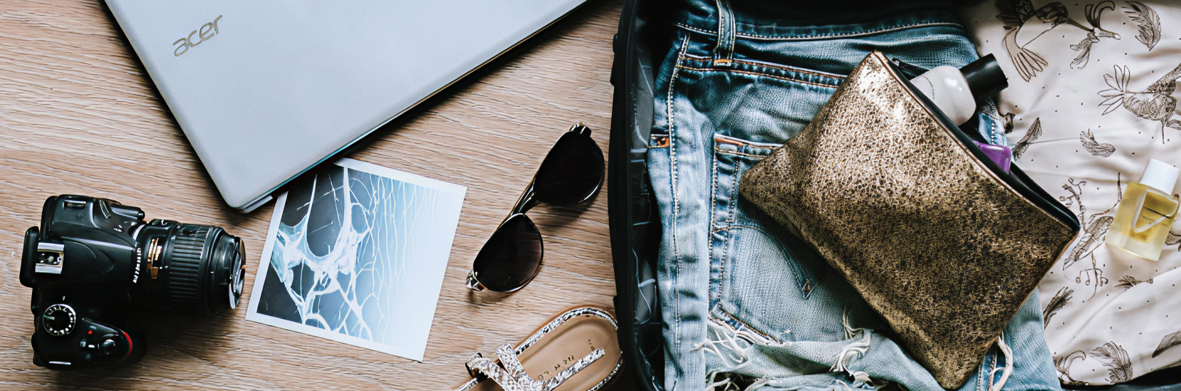 Flat lay of travel essentials including a laptop, camera, sunglasses, sandals, denim shorts, a gold makeup bag, toiletries, and a printed photo on a wooden surface
