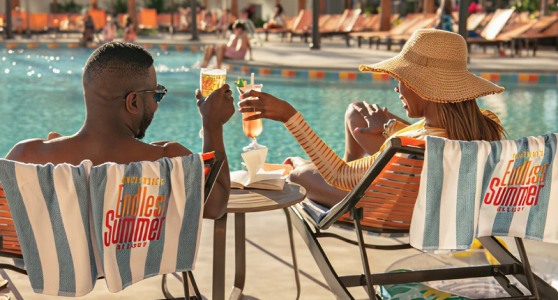 Two people clinking cocktails while reclining on poolside loungers draped with 