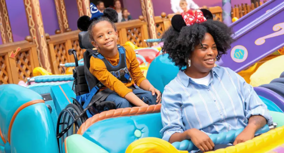 A woman wearing Minnie Mouse ears and a smiling girl in a wheelchair wearing Mickey ears enjoy a colourful theme-park ride together.