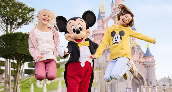 Two children jumping and smiling beside Mickey Mouse in front of a Disney castle.