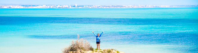 Person stading on a rock with her hands in the air. The blue sea behinde her.