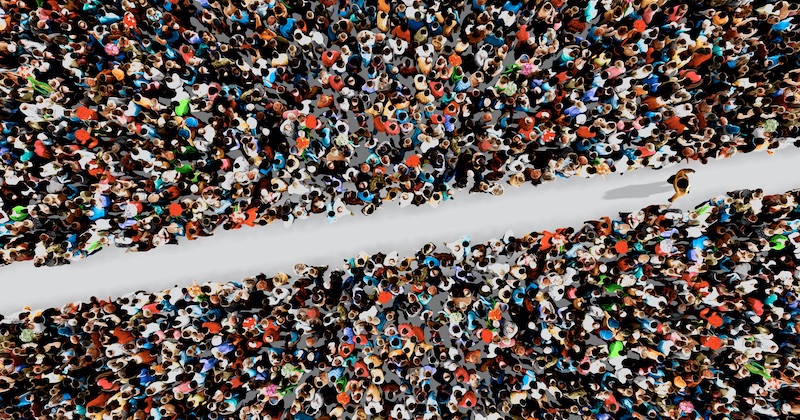 Overhead view of a dense crowd of people packed on both sides of a white walkway, colourful clothing, busy public event, high crowd density.