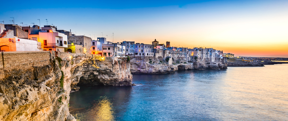 Limestone cliffs in Bari, Puglia at sunset, white buildings glowing above calm blue sea, warm lights and colourful sky – scenic Mediterranean seaside view.