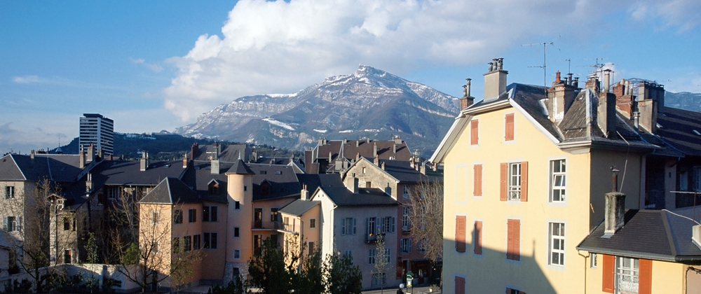 Colourful old town buildings in Chambéry, France with snow-dusted mountains in the background under a partly cloudy sky – scenic alpine city view.