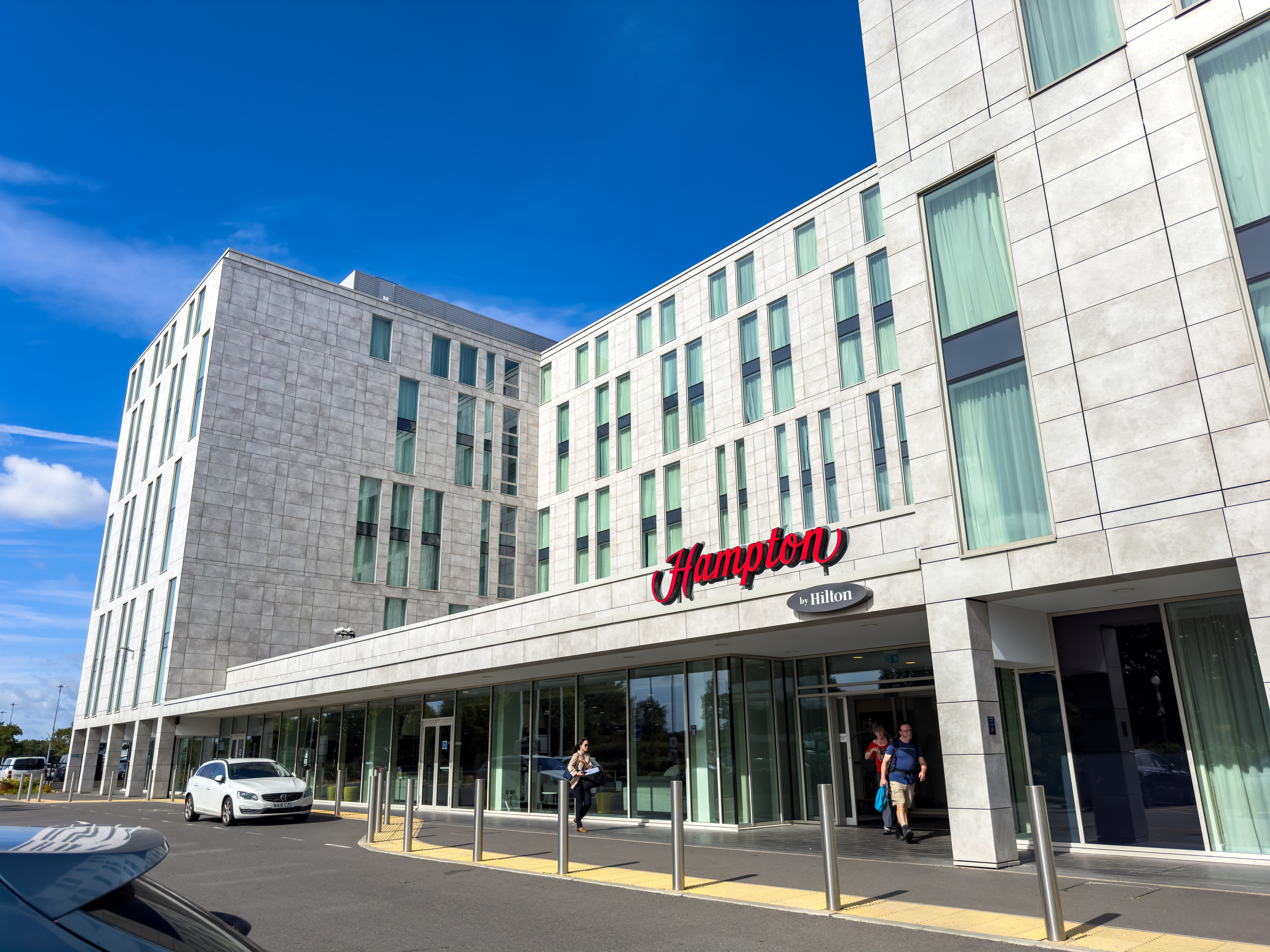 Modern Hampton by Hilton hotel with glass entrance, white facade, and vertical windows; people walking outside on a sunny day.