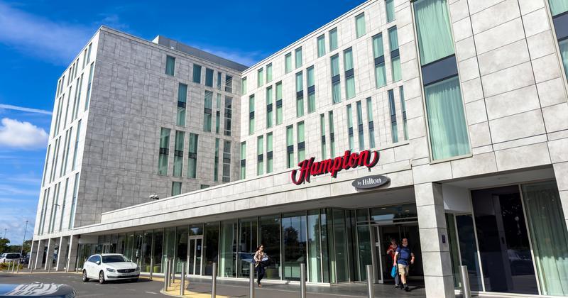 Modern Hampton by Hilton hotel with glass entrance, white facade, and vertical windows; people walking outside on a sunny day.