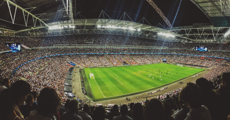 Wide-angle view of a packed football stadium at night, with thousands of spectators filling the stands around a brightly lit pitch. Players are spread across the field during a match, while floodlights illuminate the arena and beams of light shine into the night sky above.