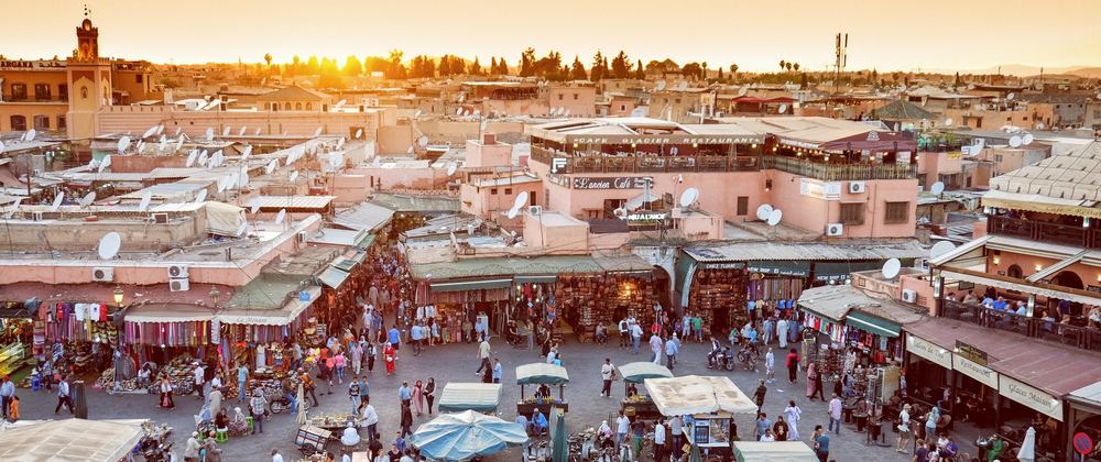 Bustling Marrakech market square at sunset with crowds shopping, rooftop cafés, pink buildings, and satellite-dotted rooftops – vibrant Moroccan cityscape.
