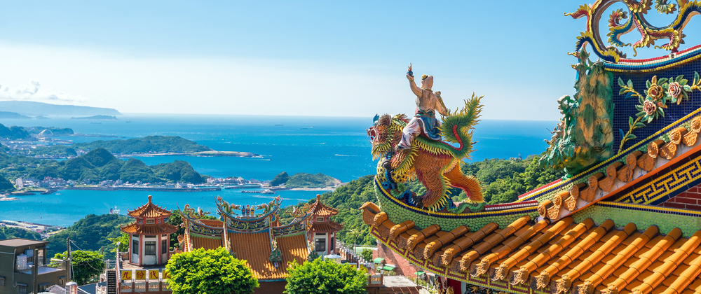 Colourful temple roof with ornate dragon statue overlooking coastal bay, blue ocean, islands and mountains – scenic Asian temple landscape.
