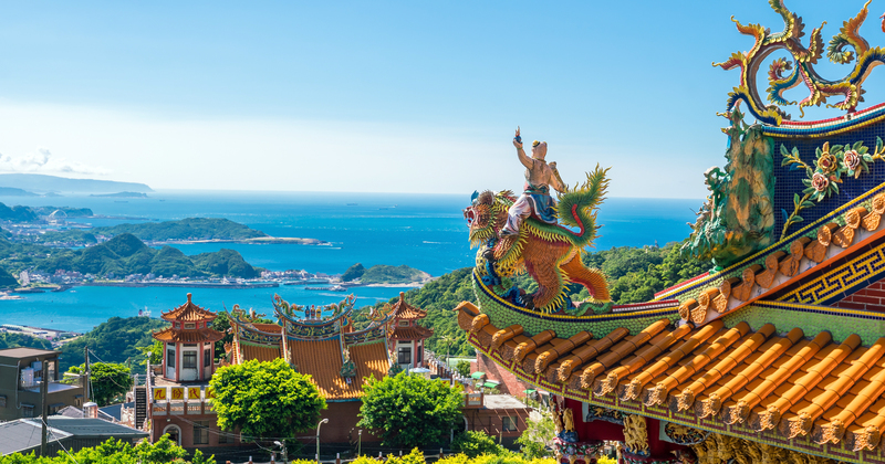 Colourful temple roof with ornate dragon statue overlooking coastal bay, blue ocean, islands and mountains – scenic Asian temple landscape.