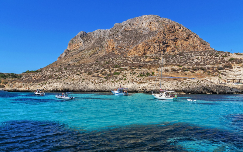 Turquoise waters and boats off the coast of Favignana, Sicily, with rocky hills in the background.