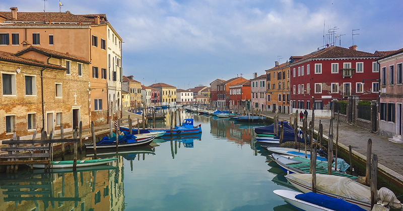 Colorful buildings and moored boats along a calm canal in Murano, Italy on a cloudy day — peaceful Venetian island known for glassmaking.