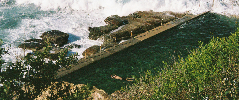 Aerial view of a coastal ocean rock pool with a swimmer floating in clear green water as waves crash against rocky shoreline — Avalon Beach, Sydney.