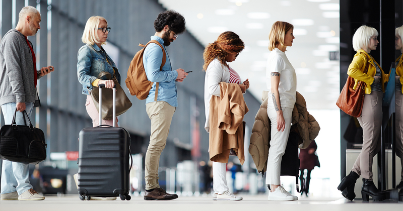 Diverse group of travelers standing in line at an airport terminal, holding luggage and coats, waiting calmly near check-in or boarding area.