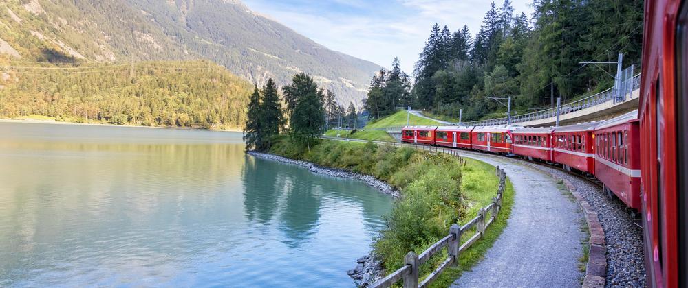 The Bernina Express, a red scenic train curving along a turquoise alpine lake with forested mountains in the background – peaceful Swiss mountain travel experience.