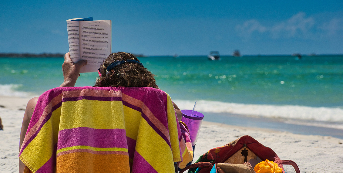Person reading a book on a beach chair covered with a colorful towel, facing turquoise ocean water with boats under a sunny blue sky — tropical beach relaxation.