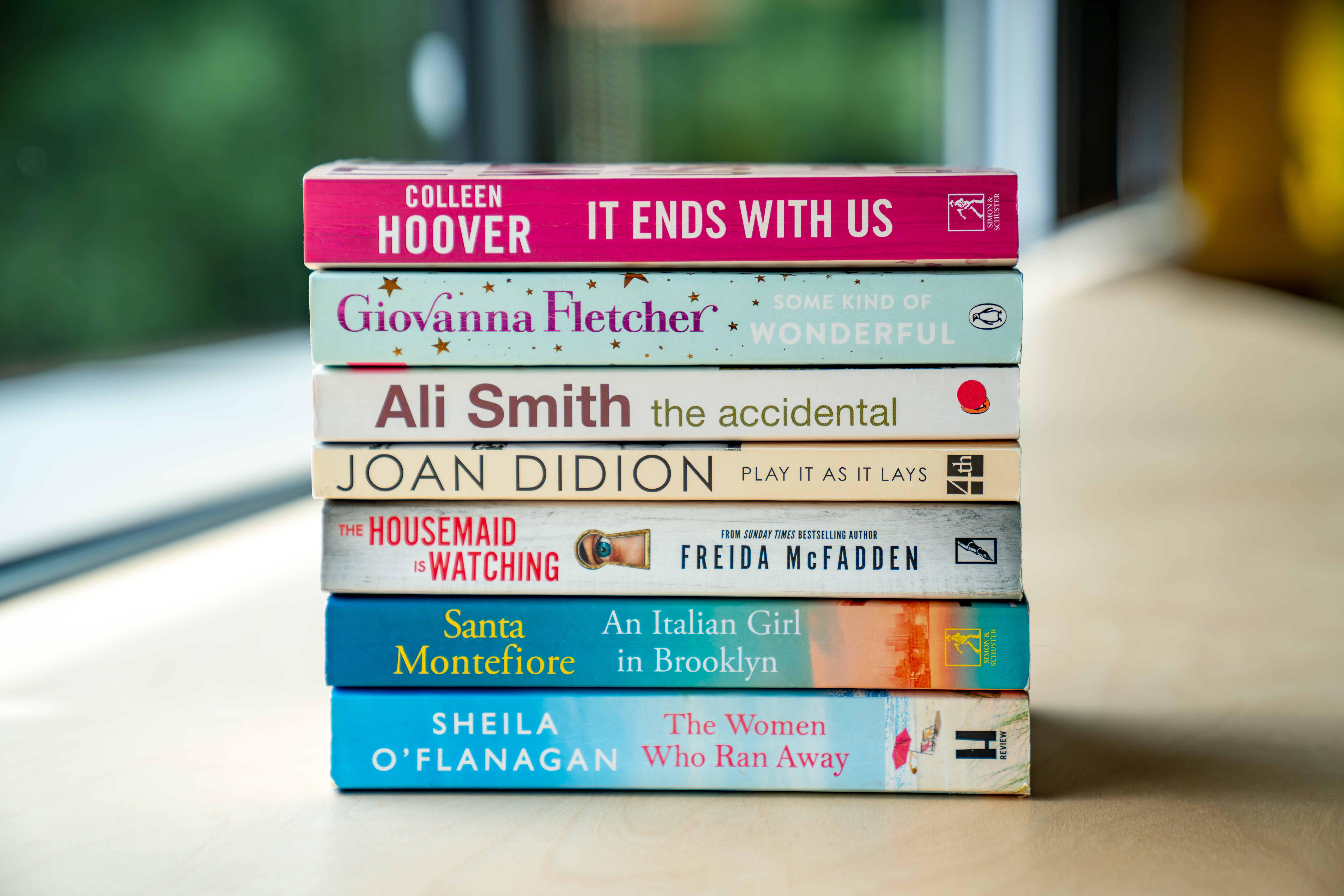 Stack of eight fiction books by popular authors including Colleen Hoover, Giovanna Fletcher, and Joan Didion, arranged on a light wooden table near a window.