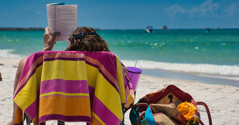 Person reading a book on a beach chair covered with a colorful towel, facing turquoise ocean water with boats under a sunny blue sky — tropical beach relaxation.