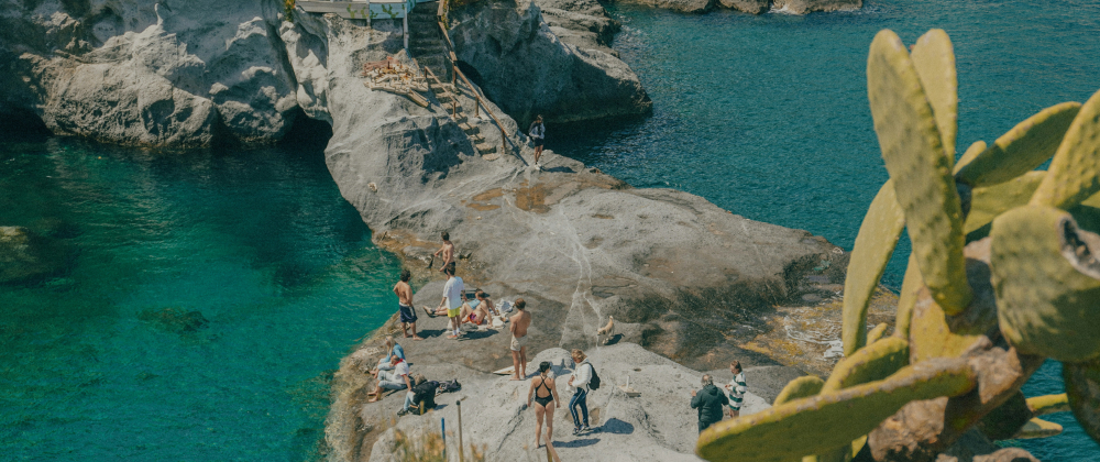 Tourists sunbathing and exploring rocky cliffs beside clear turquoise water on a Mediterranean coastline, with cactus plants in the foreground.