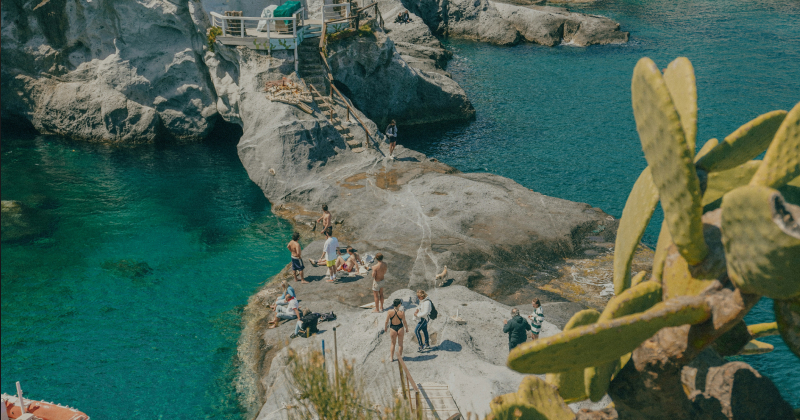 Tourists sunbathing and exploring rocky cliffs beside clear turquoise water on a Mediterranean coastline, with cactus plants in the foreground.