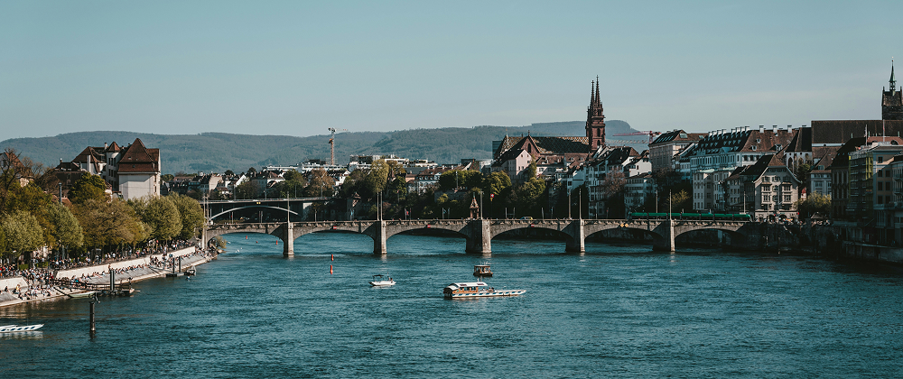 Scenic view of Basel's historic city center with the Rhine River, medieval bridge, and boats under a clear blue sky — peaceful Swiss travel destination.