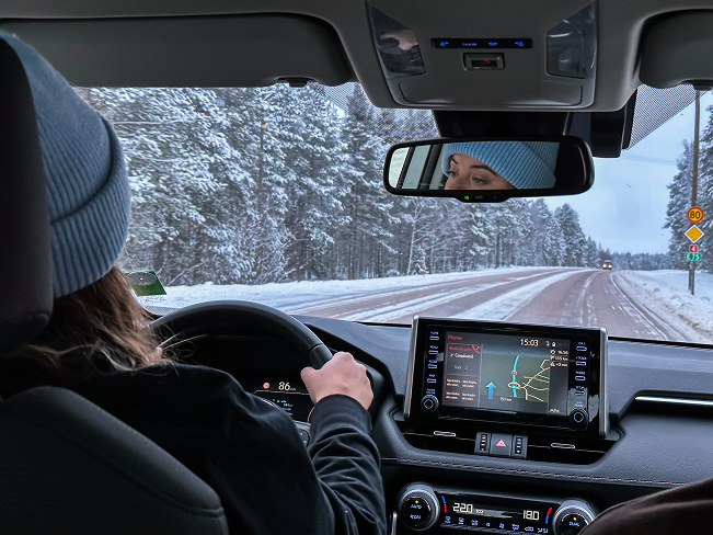 Person driving on a snowy forest road, seen from inside a modern car with GPS navigation on screen — winter road trip through scenic countryside.