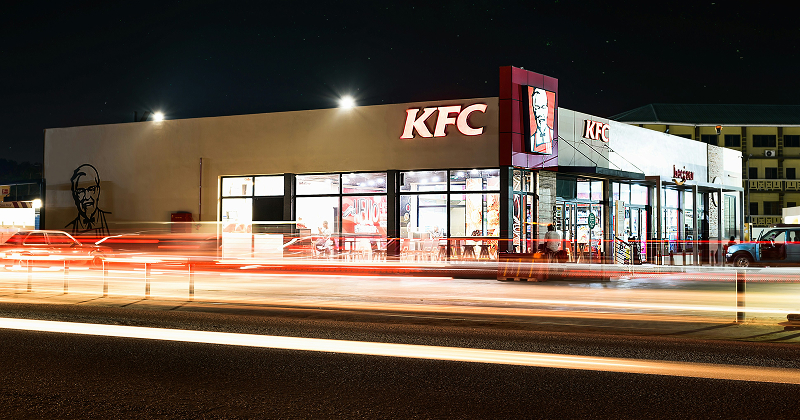 KFC fast food restaurant at night with bright lights, glowing signage, and motion blur from passing traffic — busy nighttime scene at a popular dining spot.