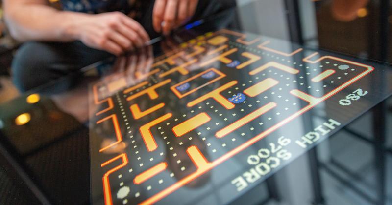 Close-up of a person playing a retro Pac-Man arcade game on a glass tabletop screen, with bright neon maze and high score display — classic video game nostalgia.