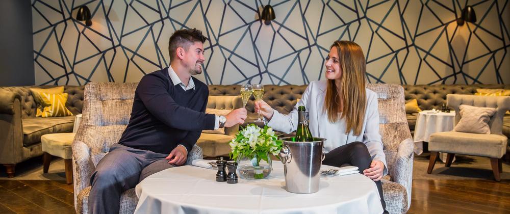 Smiling couple toasting champagne at an elegant airport lounge table with flower centerpiece and modern geometric wall decor — romantic fine dining experience.