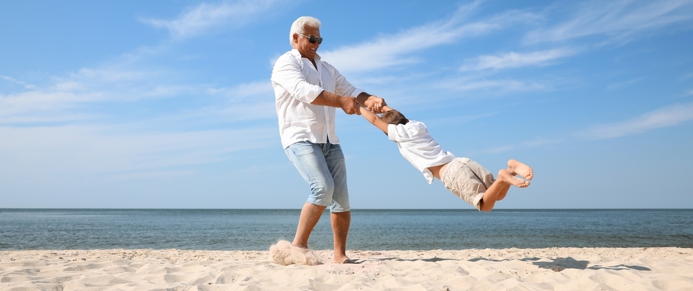 Smiling grandfather swinging young boy by the arms on a sunny beach with blue sky and calm ocean – joyful family moment by the sea.