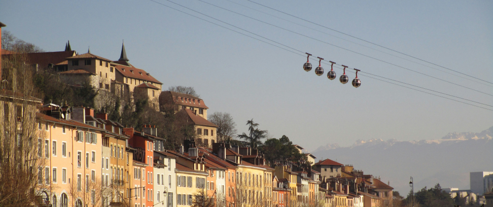 Colourful hillside houses beneath a historic fortress with cable cars overhead and distant snowy mountains – scenic European city landscape at sunset.