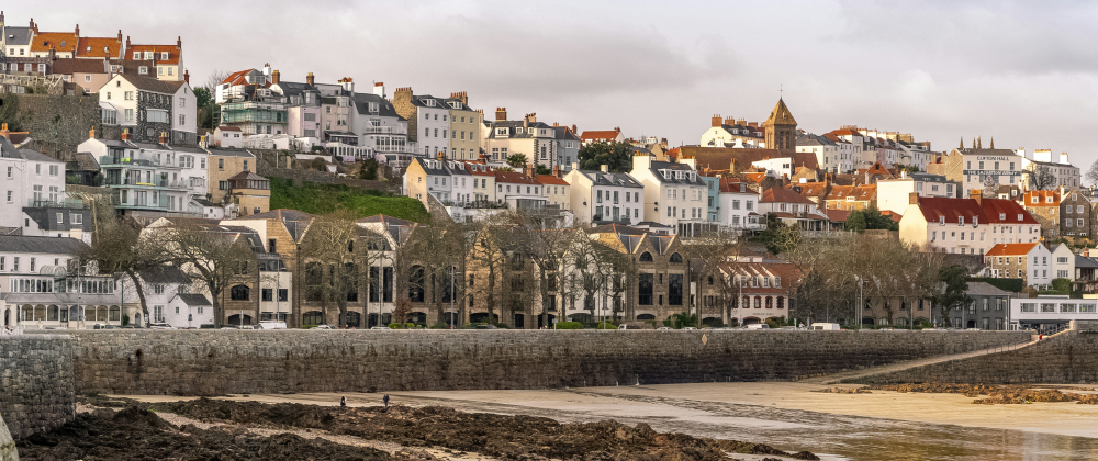 Charming seaside town in Guernsey with historic stone buildings, white cottages, and a church spire on a hillside above a stone seawall and sandy beach at low tide.