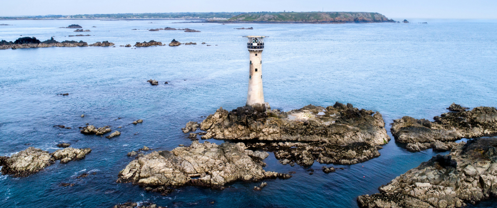 Les Hanois Lighthouse, Guernsey. A tall stone lighthouse on rocky islet surrounded by calm blue sea, with distant coastline and scattered rock formations — remote coastal navigation landmark.