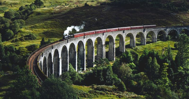 Steam train crossing the Glenfinnan Viaduct in Scotland, surrounded by lush green hills and forest — iconic scenic railway journey.