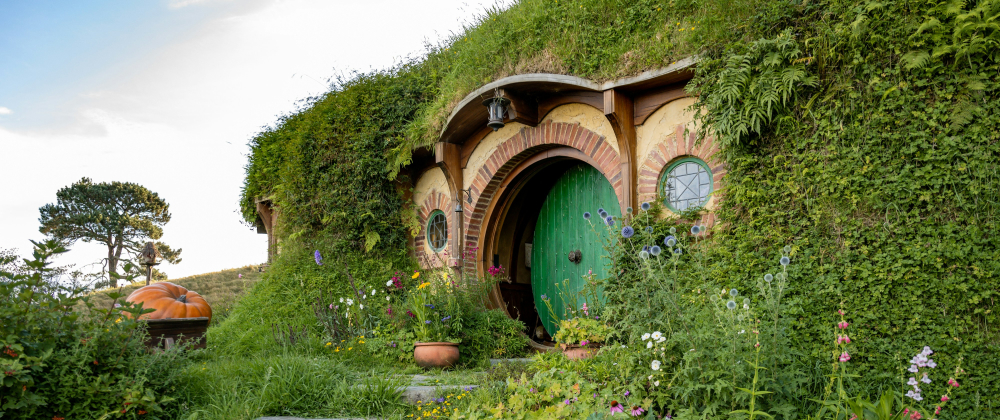 Hobbit house with round green door and circular windows built into a grassy hillside, surrounded by colourful flowers and lush greenery.