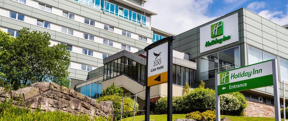 Holiday Inn hotel exterior beside Edinburgh Zoo car park entrance, modern building with green signage, landscaped grounds, and blue sky.