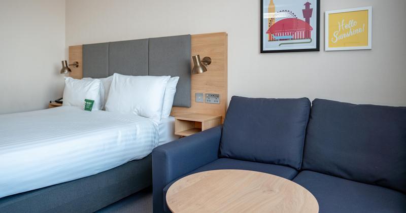 Modern hotel room at Holiday Inn Gatwick Airport with double bed, navy sofa, round coffee table, and cheerful wall art — clean, comfortable stay near airport.