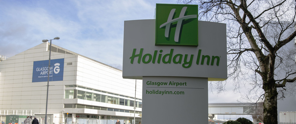 Holiday Inn Glasgow Airport sign outside the hotel, with Glasgow Airport terminal building in the background under a cloudy sky.