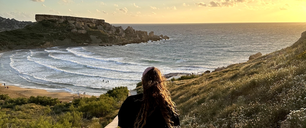 Woman overlooking Riviera Beach in Malta at sunset with rolling ocean waves, sandy shoreline, rocky cliffs, and golden light – peaceful seaside landscape.