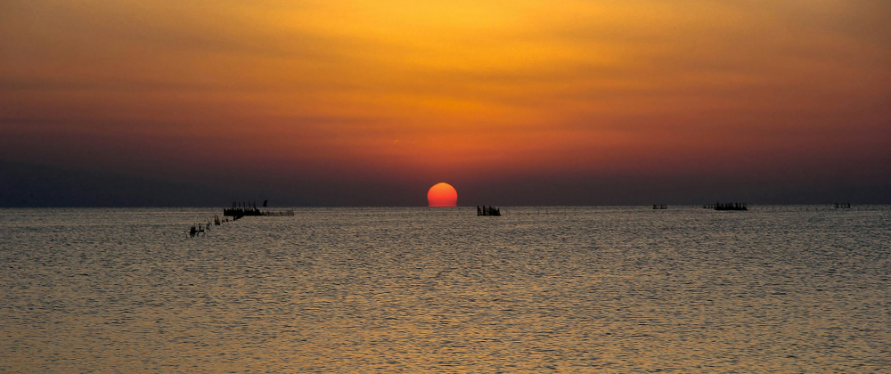 Sun setting over calm ocean horizon with orange sky, small fishing boats silhouetted on the water – serene coastal sunset landscape.