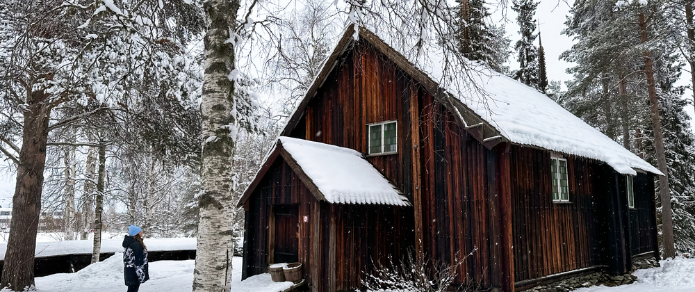 Person walking up to Sodankylä Old Church, a 17th-century wooden church near the Kitinen River in Sodankylä, Lapland. The church has a snow-covered roof and is surrounded by pine trees and winter scenery – peaceful Finnish winter landscape.